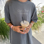 Person wearing a gray t-shirt with 'God's Still Working' text, holding an iced coffee.