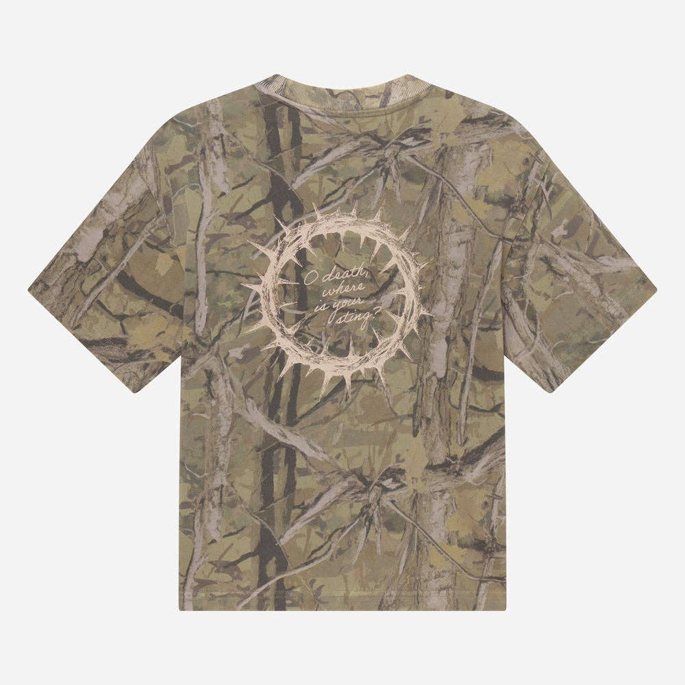 A short-sleeve t-shirt with an all-over woodland camouflage pattern in shades of brown, beige, and green. The center of the chest features a circular graphic design with a thorny crown of thorns motif and the text 'But the Lord loves righteousness' in a light beige font.
