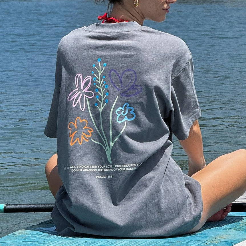 Woman sitting on a paddleboard by a body of water wearing a gray t-shirt with colorful floral designs.