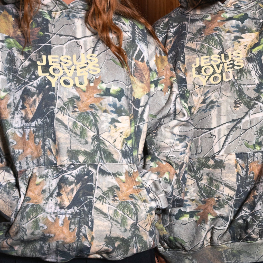 Two models wearing matching camouflage hoodies with the text 'JESUS LOVES YOU' printed in gold on the chest.