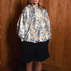 A woman stands against a wood-paneled wall wearing a camouflage-patterned hooded sweatshirt and black shorts.