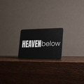 A black rectangular card or plaque with the words "HEAVEN below" in white, standing upright on a dark wooden surface against a textured taupe wall.
