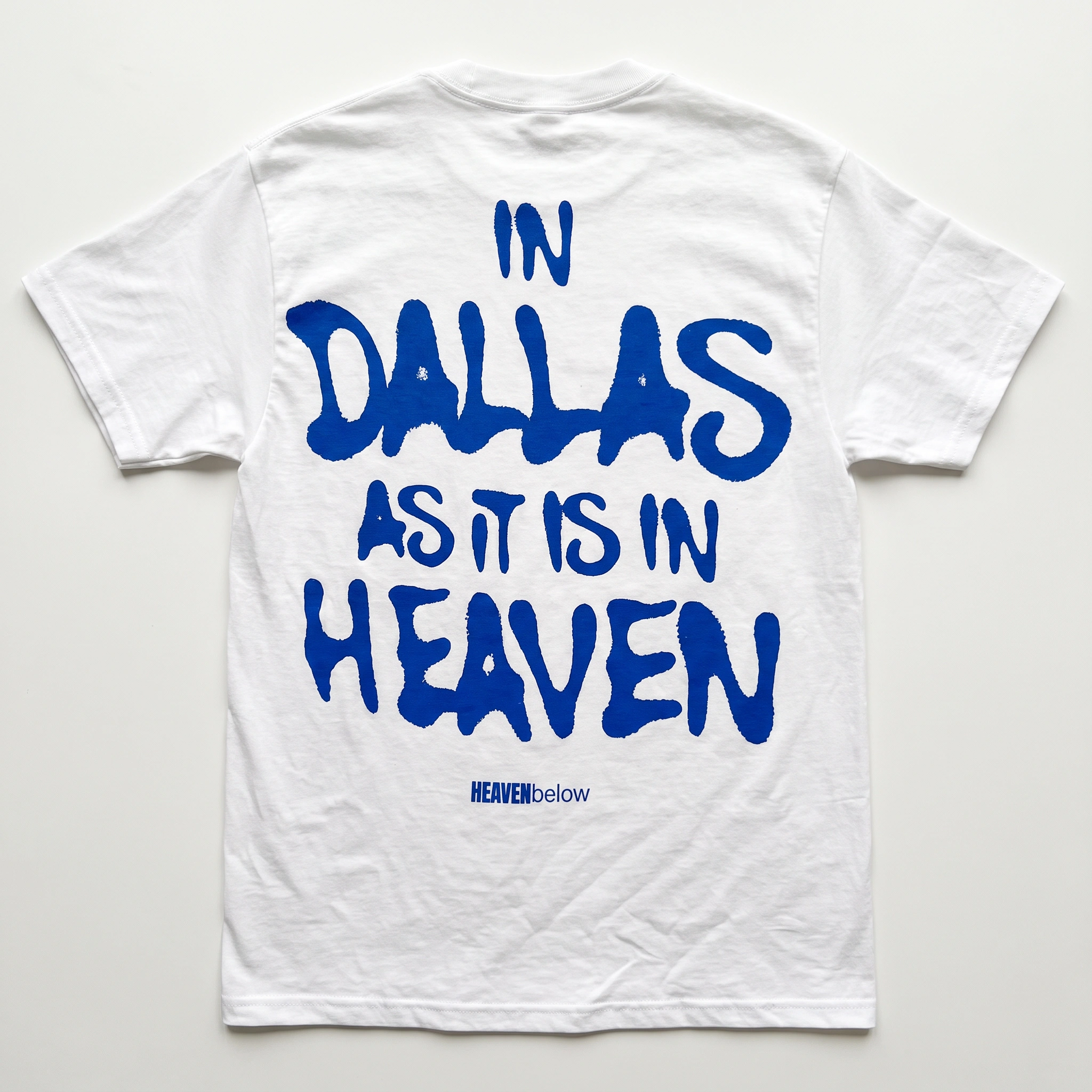 White short-sleeve t-shirt laid flat on a light background, featuring the phrase 'IN DALLAS AS IT IS IN HEAVEN' printed in large blue letters across the back, with 'HEAVEN below' in smaller blue text underneath.