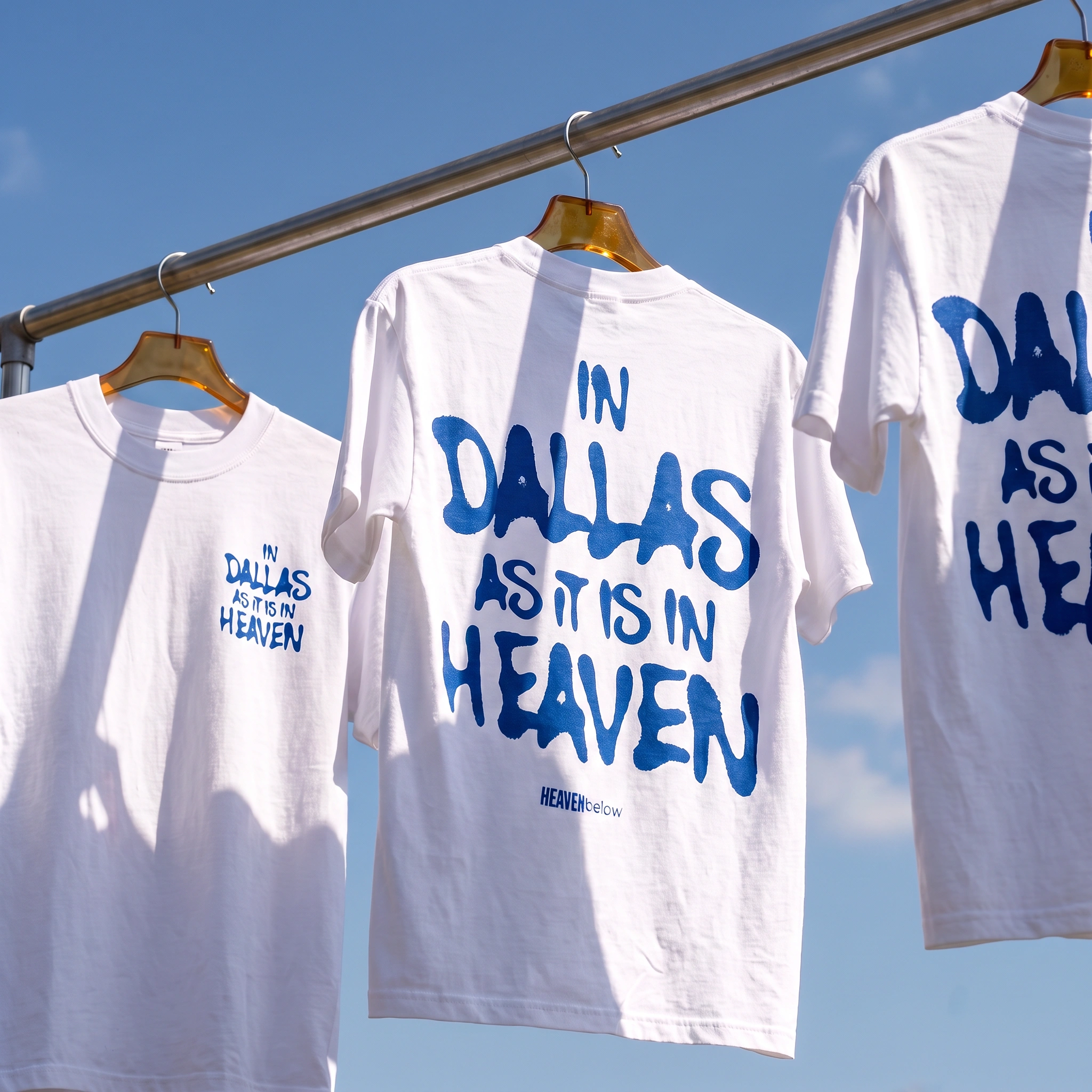Three white graphic T-shirts hanging on wooden hangers from a metal rail against a blue sky. The center shirt shows the back with large blue text reading 'IN DALLAS AS IT IS IN HEAVEN,' while the side shirts show the front with smaller matching blue text.