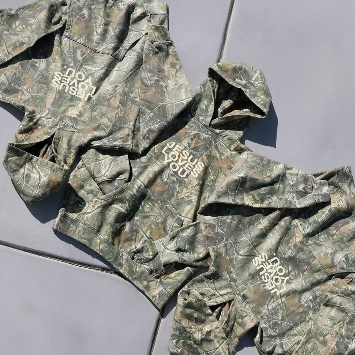 Three camouflage hoodies in green, brown, and beige tones are laid out on a gray tiled floor. Each hoodie features a white graphic text print on the chest, including 'DO NOT DISTURB' and 'JUST SHUT UP'.