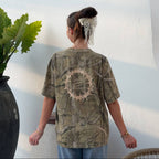 A person viewed from behind wearing an oversized short-sleeve t-shirt with a distressed camouflage pattern in olive green, tan, and brown, featuring a circular graphic print on the back. The background is a clean white wall with a decorative scalloped awning and a potted plant.