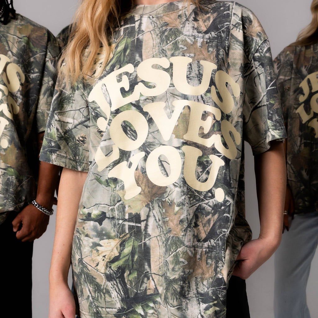 Person wearing a camouflage t-shirt with 'Jesus Loves You' text.