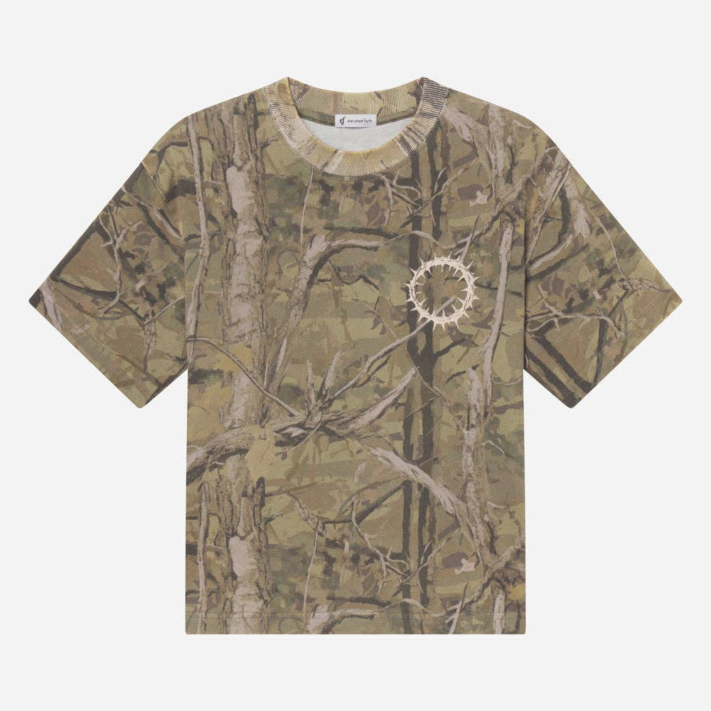 A short-sleeve crewneck t-shirt featuring an all-over woodland camouflage pattern with branches and leaves in shades of brown, beige, and green. A small white circular graphic is printed on the left chest.