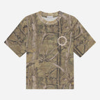 A short-sleeve crewneck t-shirt featuring an all-over woodland camouflage pattern with branches and leaves in shades of brown, beige, and green. A small white circular graphic is printed on the left chest.