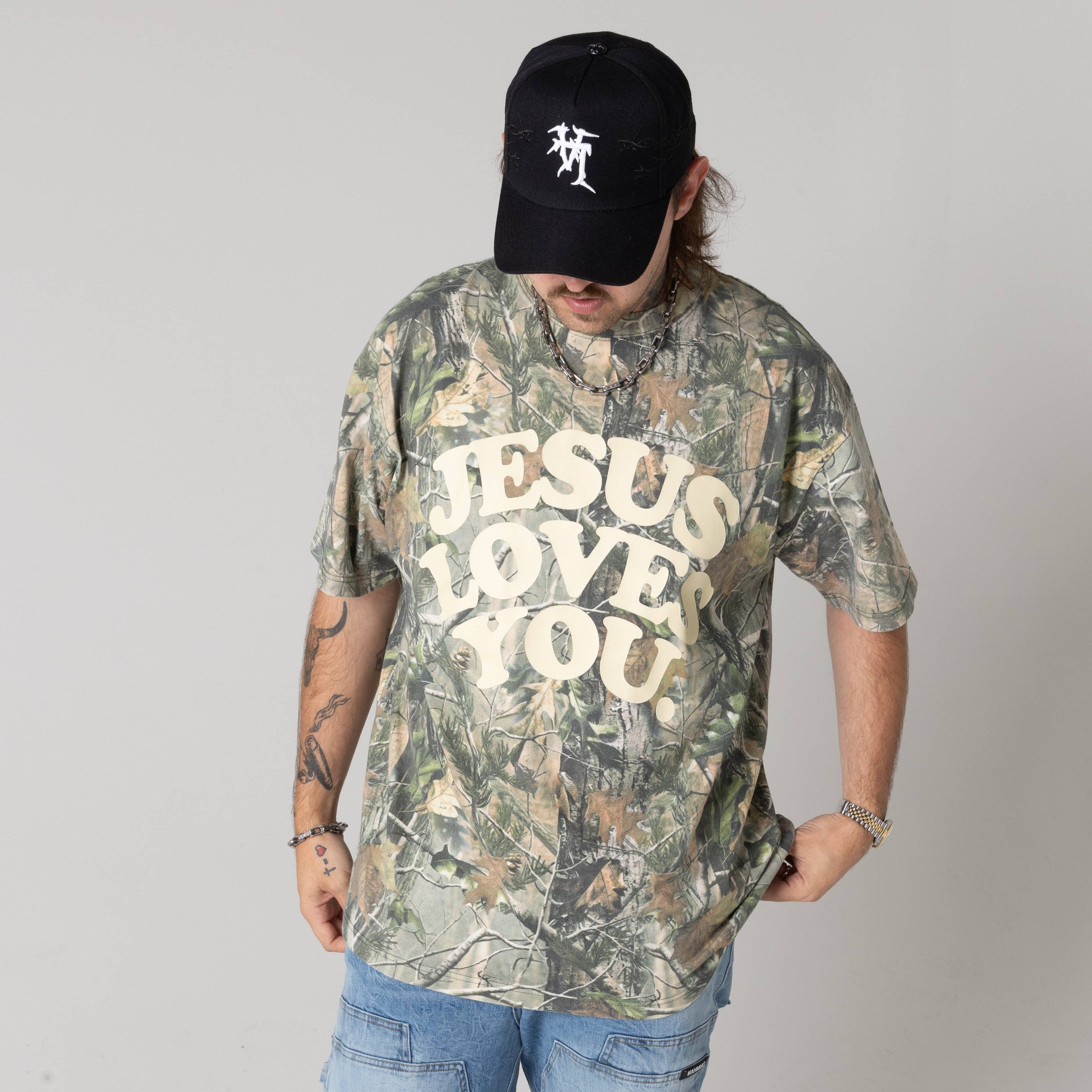 Person wearing a camouflage t-shirt with 'Jesus Loves You' text and a black cap.